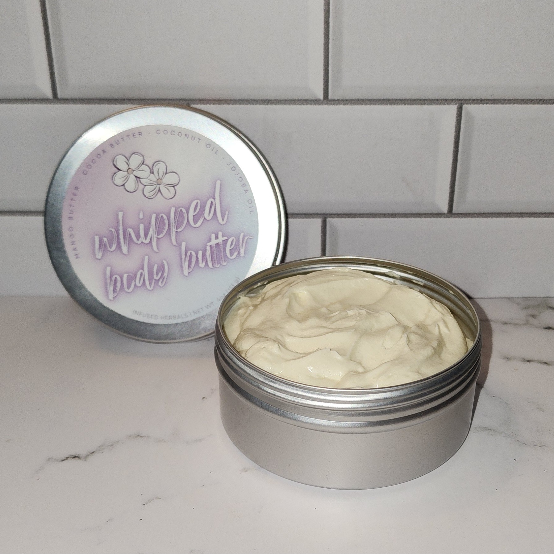 A 4oz tin jar of whipped body butter with a sliver screw on lid with a lavender and blue gradient product label.