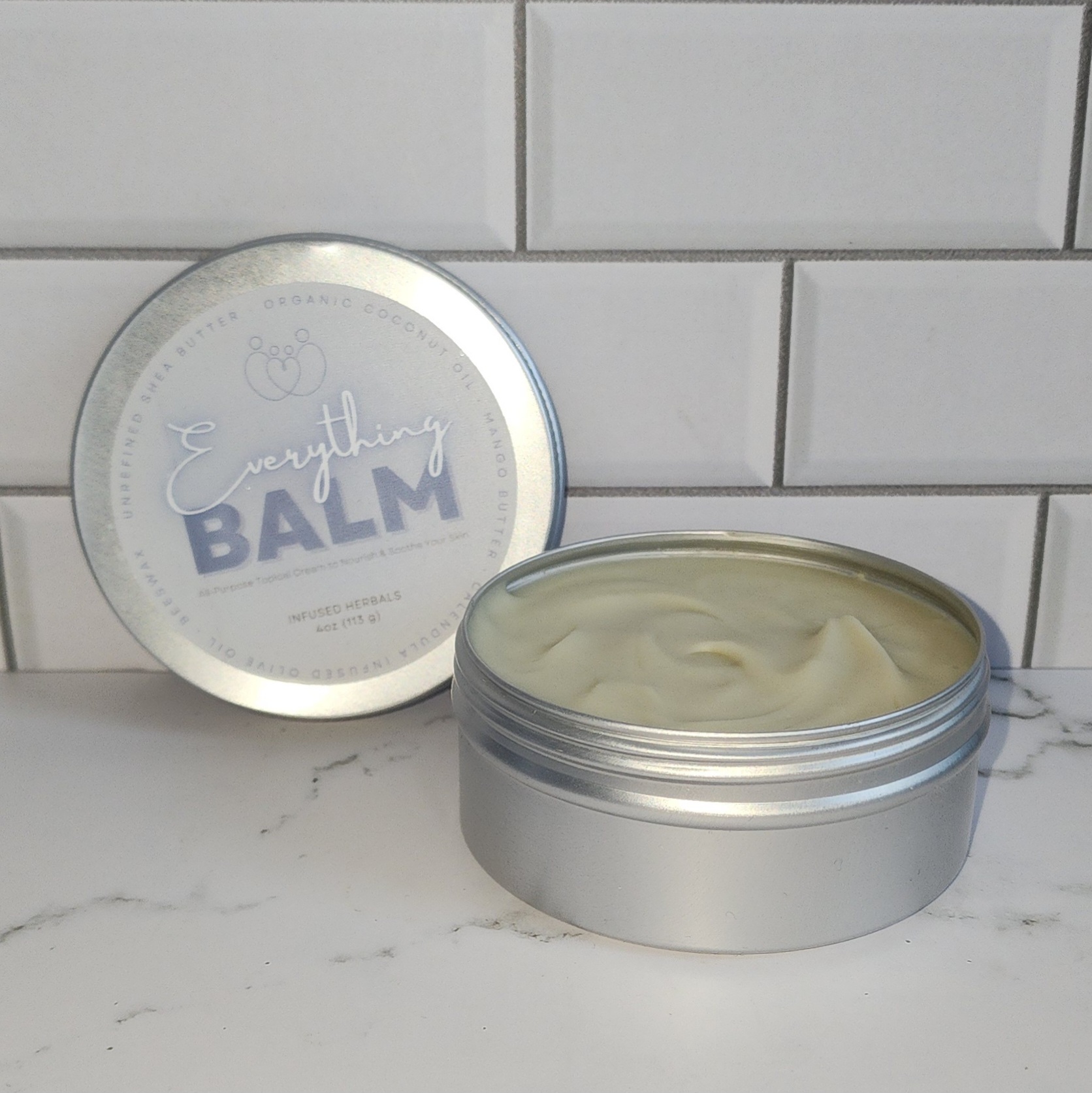 A 4oz tin of a homemade balm set on a marble countertop in front of a white tile backsplash.